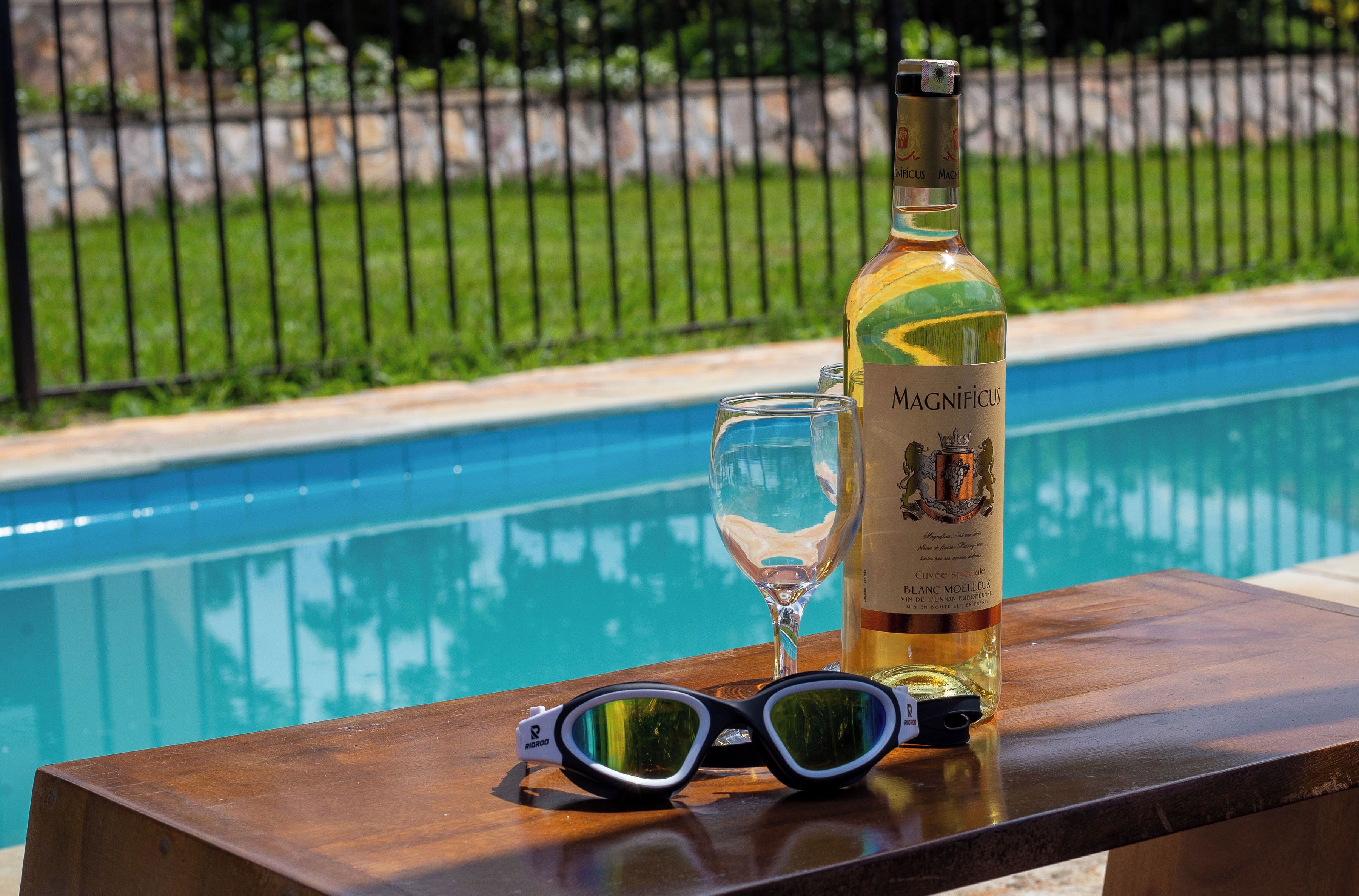 Uma garrafa de vinho Magnificus, uma taça de vinho e um par de óculos de sol sobre uma mesa de madeira junto a uma piscina.