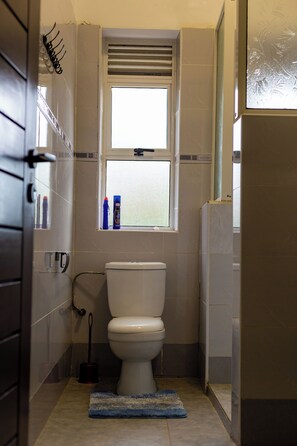 A bathroom with a toilet, a window, and a shower area. - Martha's Farm House (Mukono)