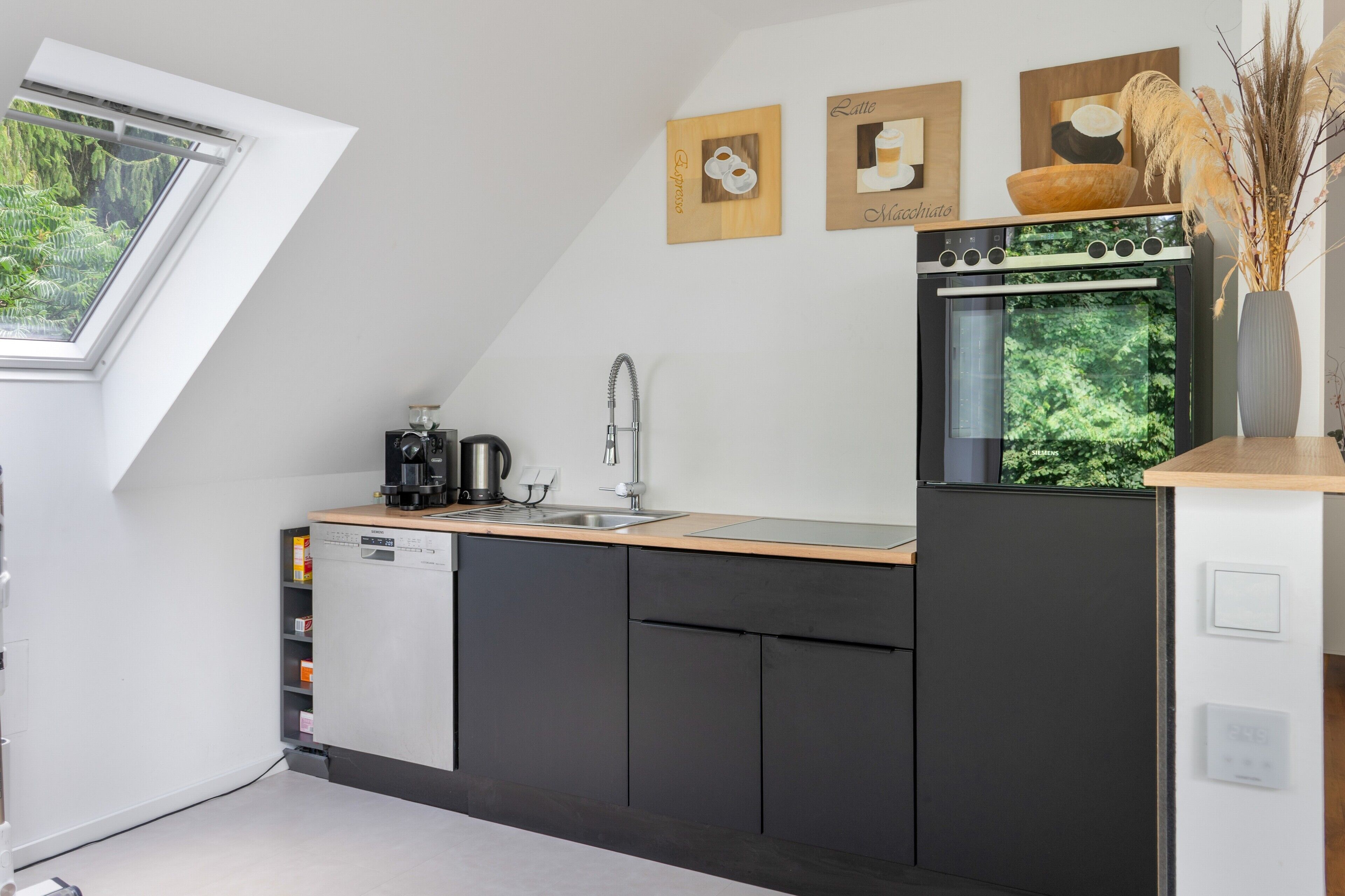 A modern kitchen with a built-in dishwasher, a coffee machine, and a sink. There are framed pictures on the wall and a window with a view of greenery.