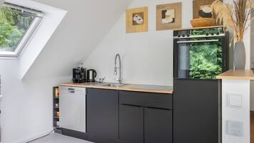 A modern kitchen with a built-in dishwasher, a coffee machine, and a sink. There are framed pictures on the wall and a window with a view of greenery.