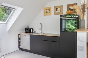 A modern kitchen with a built-in dishwasher, a coffee machine, and a sink. There are framed pictures on the wall and a window with a view of greenery. - Stayplace No. 1: Open-Plan Design Apartment with Two Bedrooms and Modern Amenities (Garstedt)