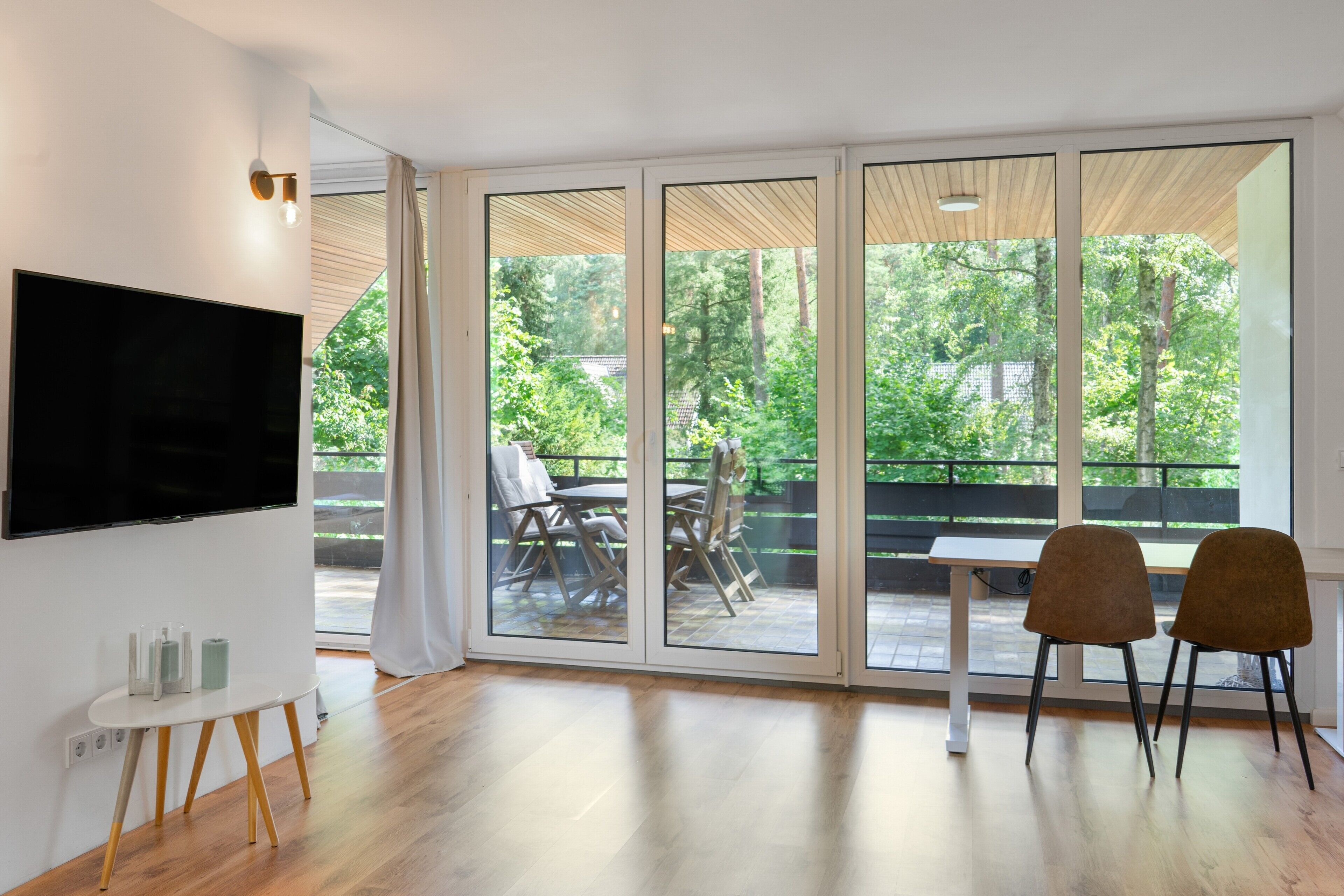 Ein modernes Wohnzimmer mit Flachbildfernseher, einem Esstisch mit Stühlen und einer Schiebetür zu einer Terrasse im Freien mit Blick auf Bäume.