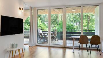 Ein modernes Wohnzimmer mit Flachbildfernseher, einem Esstisch mit Stühlen und einer Schiebetür zu einer Terrasse im Freien mit Blick auf Bäume.