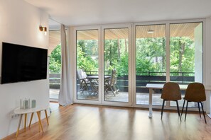 A modern living room with a flat-screen TV, a dining table with chairs, and a sliding glass door leading to an outdoor patio with a view of trees. - Stayplace No. 1: Open-Plan Design Apartment with Two Bedrooms and Modern Amenities (Garstedt)