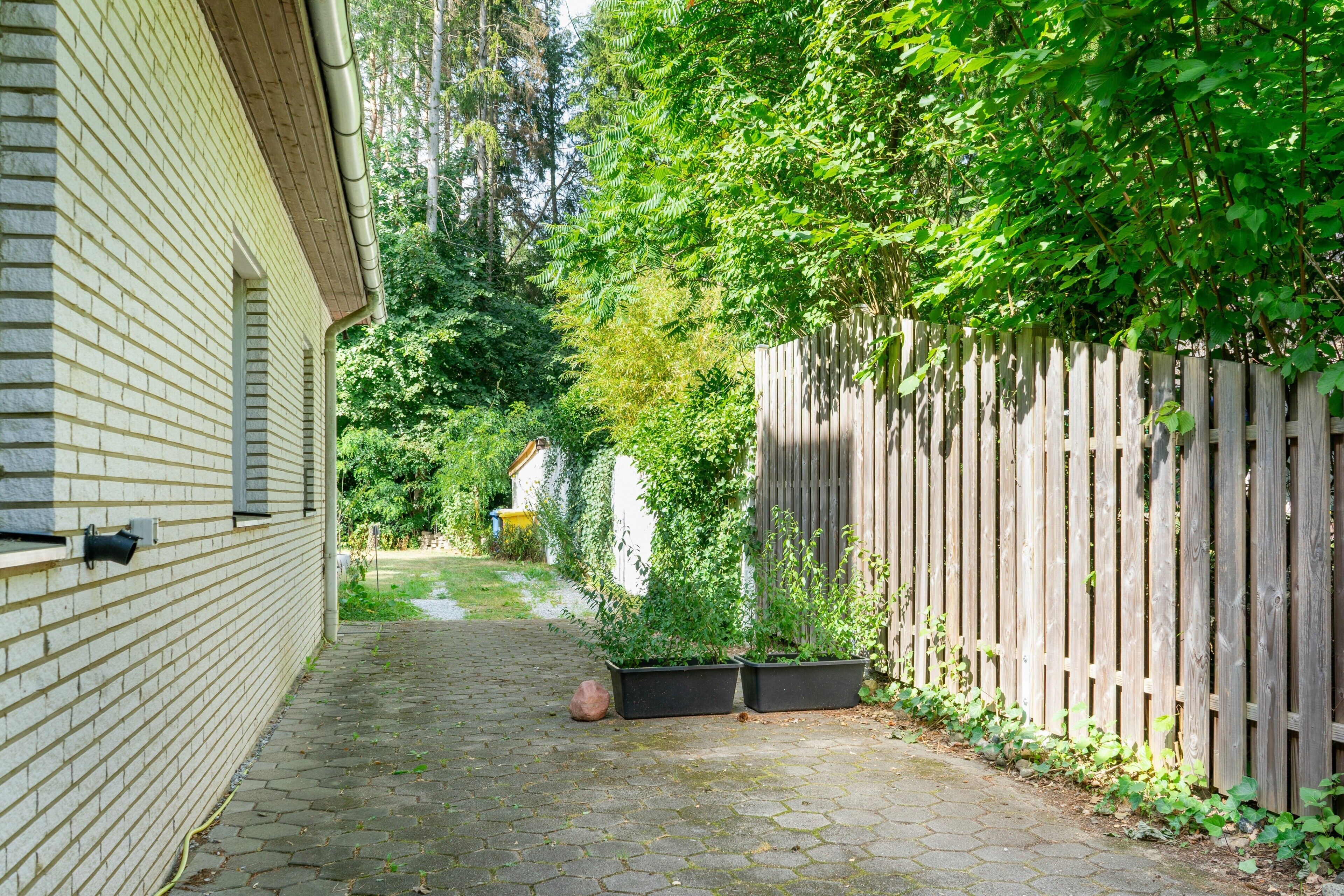 Ein schmaler, gepflasterter Weg, der von einer Backsteinmauer und einem Holzzaun gesäumt wird, führt zu einem Gartenbereich mit Kübelpflanzen und einem kleinen Schuppen.