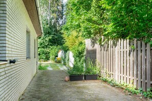Un estrecho sendero empedrado flanqueado por un muro de ladrillo y una valla de madera, que conduce a un jardín con plantas en macetas y un pequeño cobertizo.