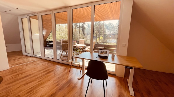 A modern room with a wooden floor, a desk with a laptop, a chair, and a view through large glass doors to an outdoor area with a table and chairs.