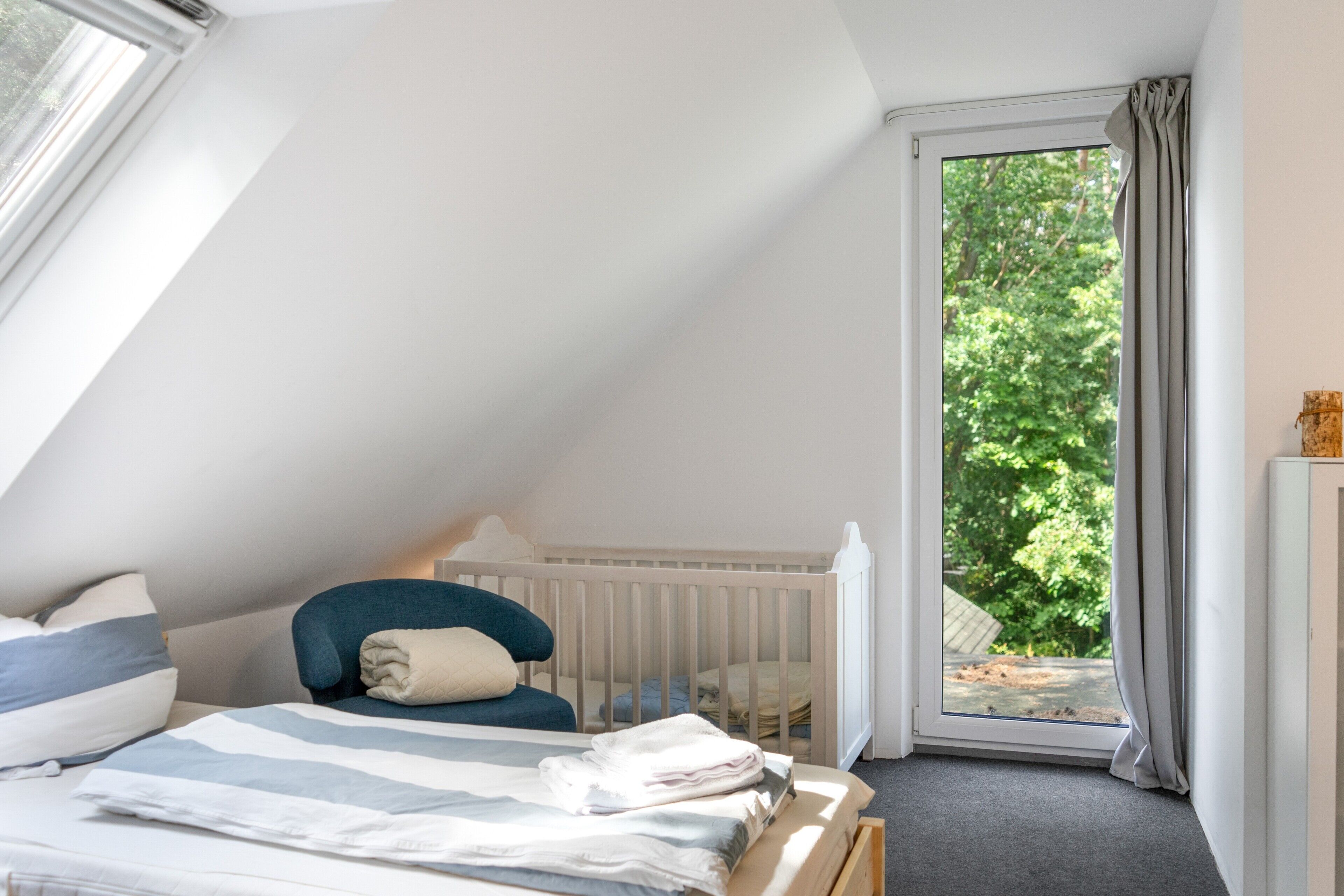 A bedroom with a sloped ceiling, a bed with a striped bedspread, a chair, a crib, and a view of greenery through a window.
