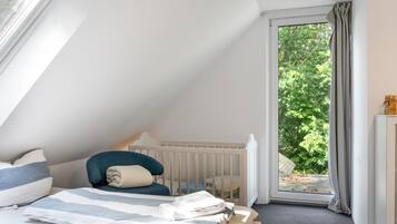 A bedroom with a sloped ceiling, a bed with a striped bedspread, a chair, a crib, and a view of greenery through a window.