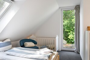 Un dormitorio con techo inclinado, una cama con colcha a rayas, una silla, una cuna y vista a la vegetación a través de una ventana.