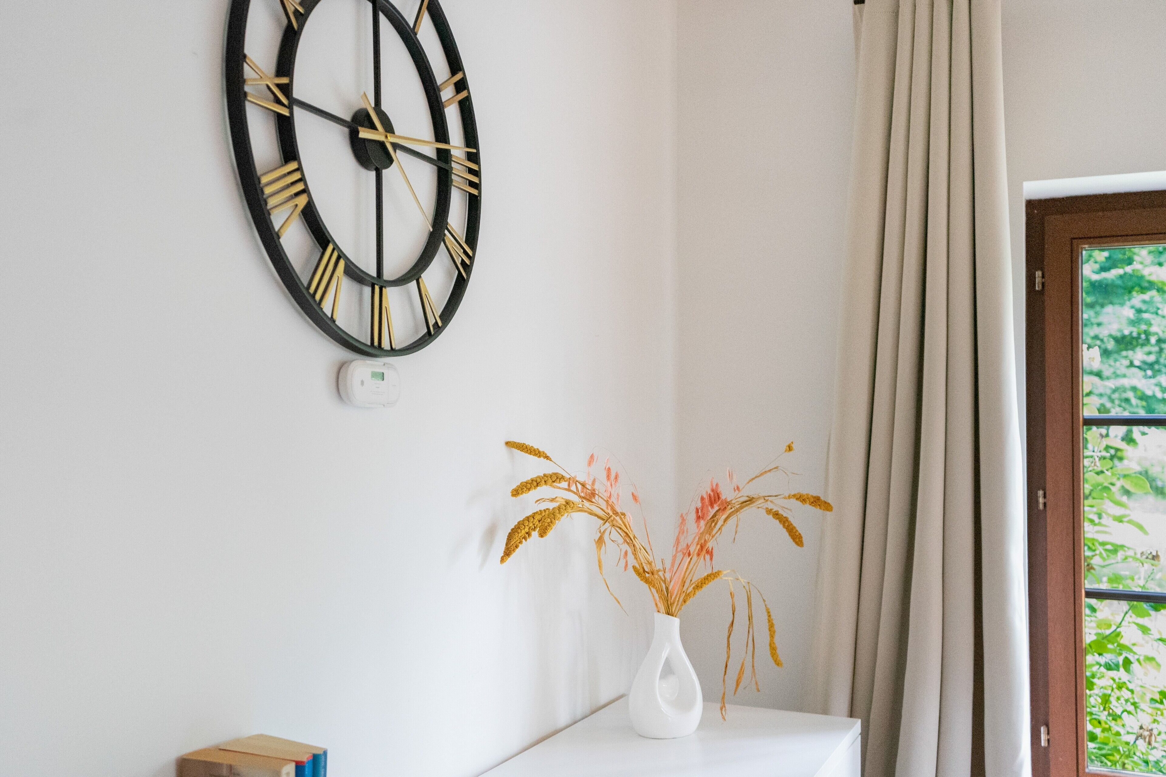 A room with a wall-mounted clock, a white vase with dried plants, and a window with curtains.
