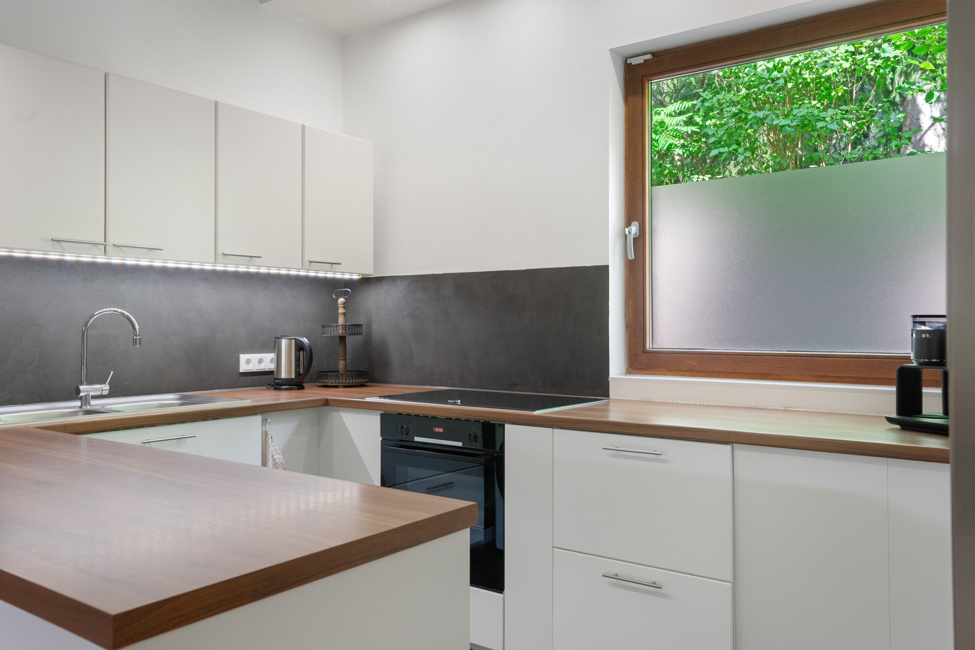 A modern kitchen with white cabinets, a wooden countertop, a built-in oven, and a window with a view of greenery.