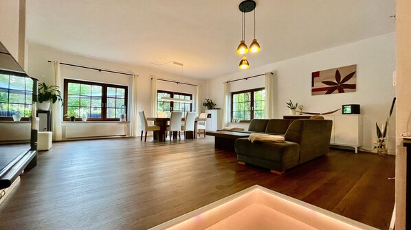 A spacious living area with a dining table, sofa, and large windows.