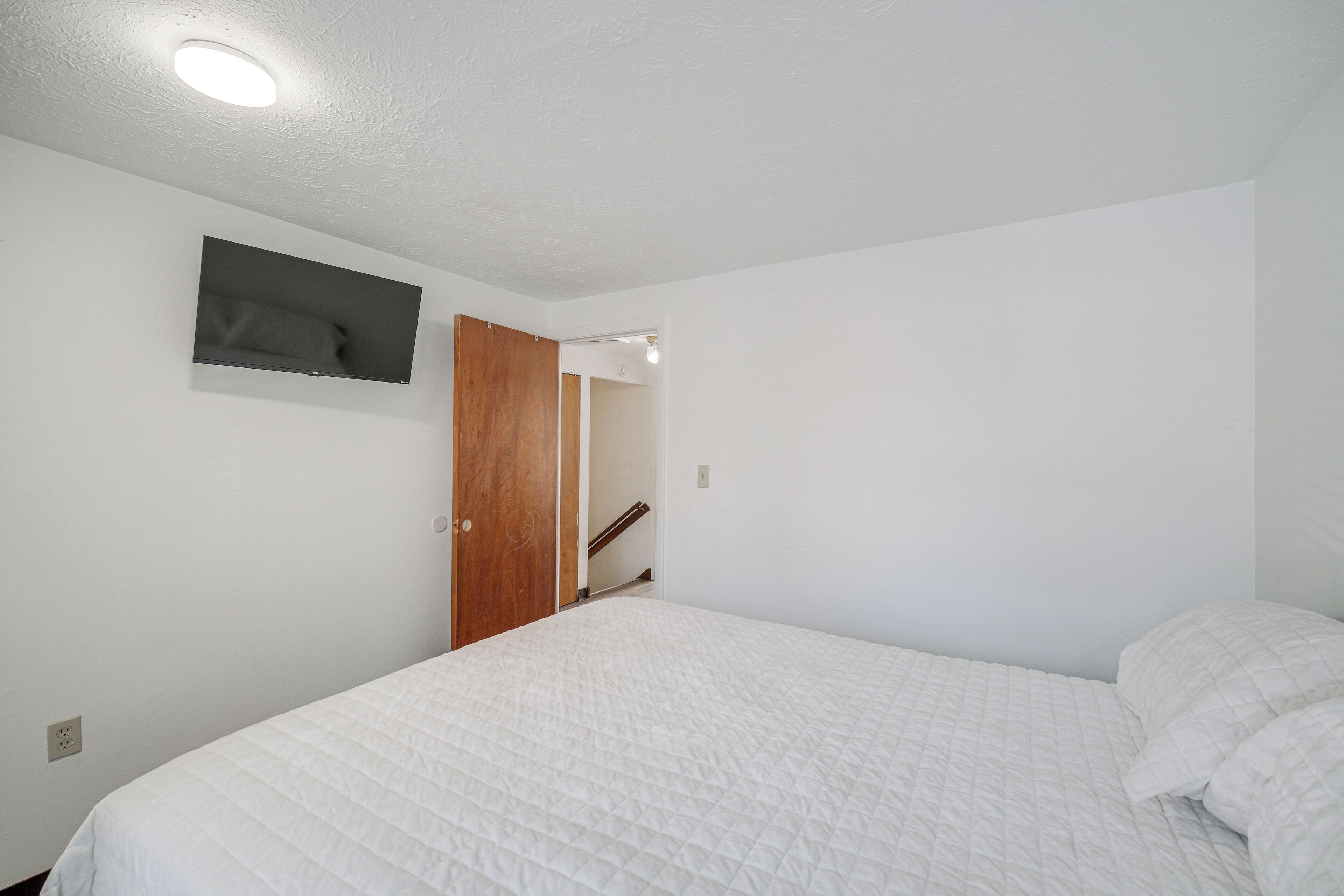 A bedroom with a bed, a TV mounted on the wall, and a wooden door leading to another room.