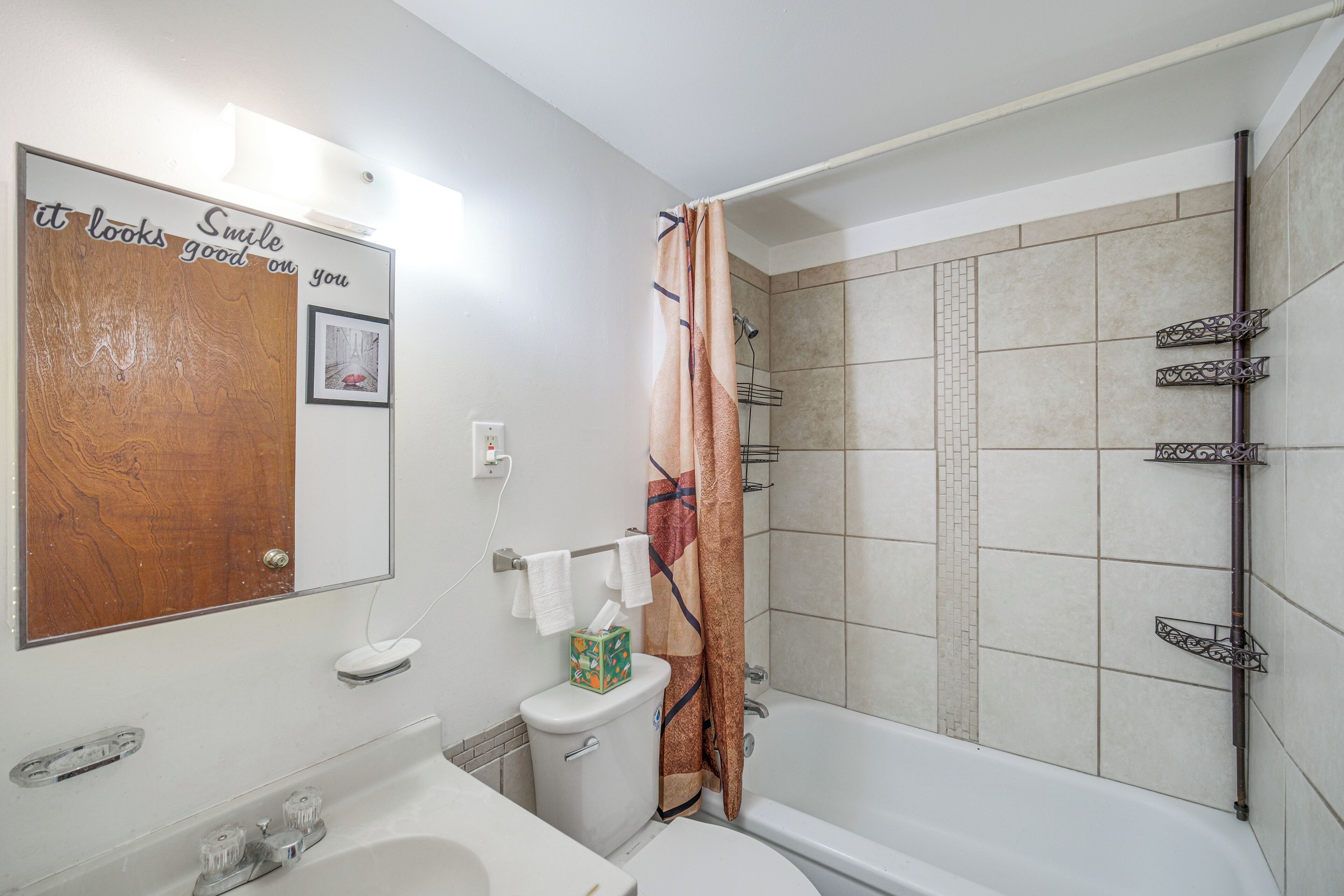 Un baño con puerta de madera, un lavamanos blanco, inodoro, ducha con cortina estampada y bañera.