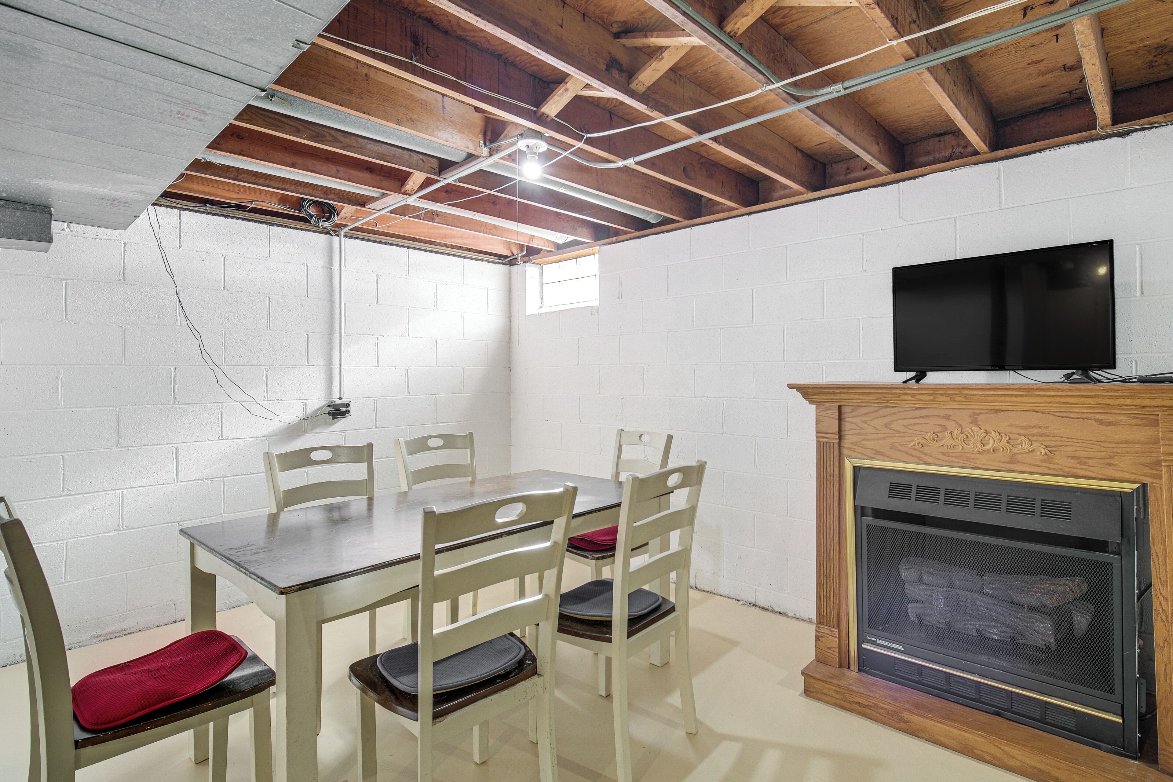A room with a wooden ceiling, white brick walls, a TV mounted above a fireplace, and a dining table with chairs.