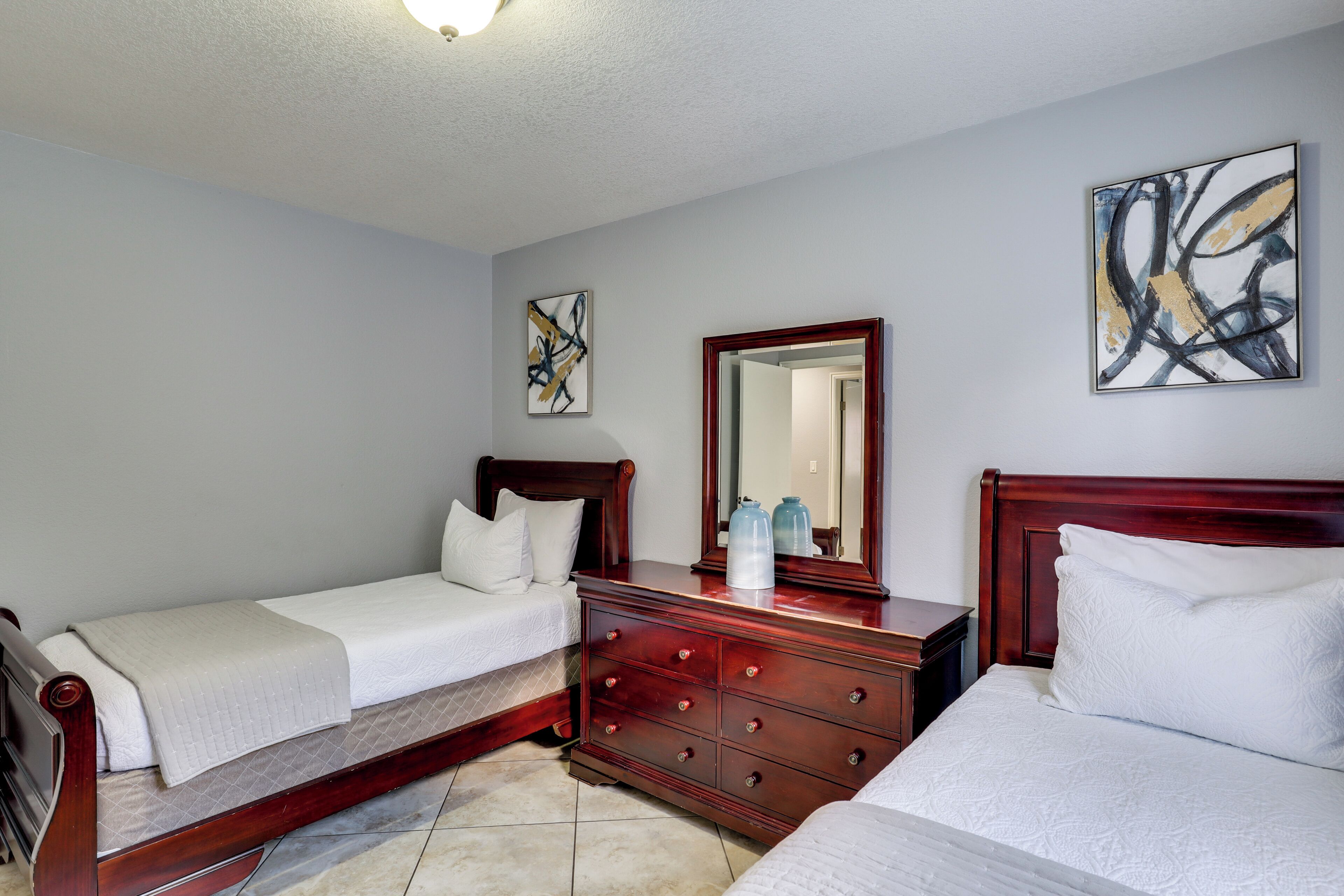 A hotel room with two single beds, a wooden dresser, a mirror, and abstract wall art.