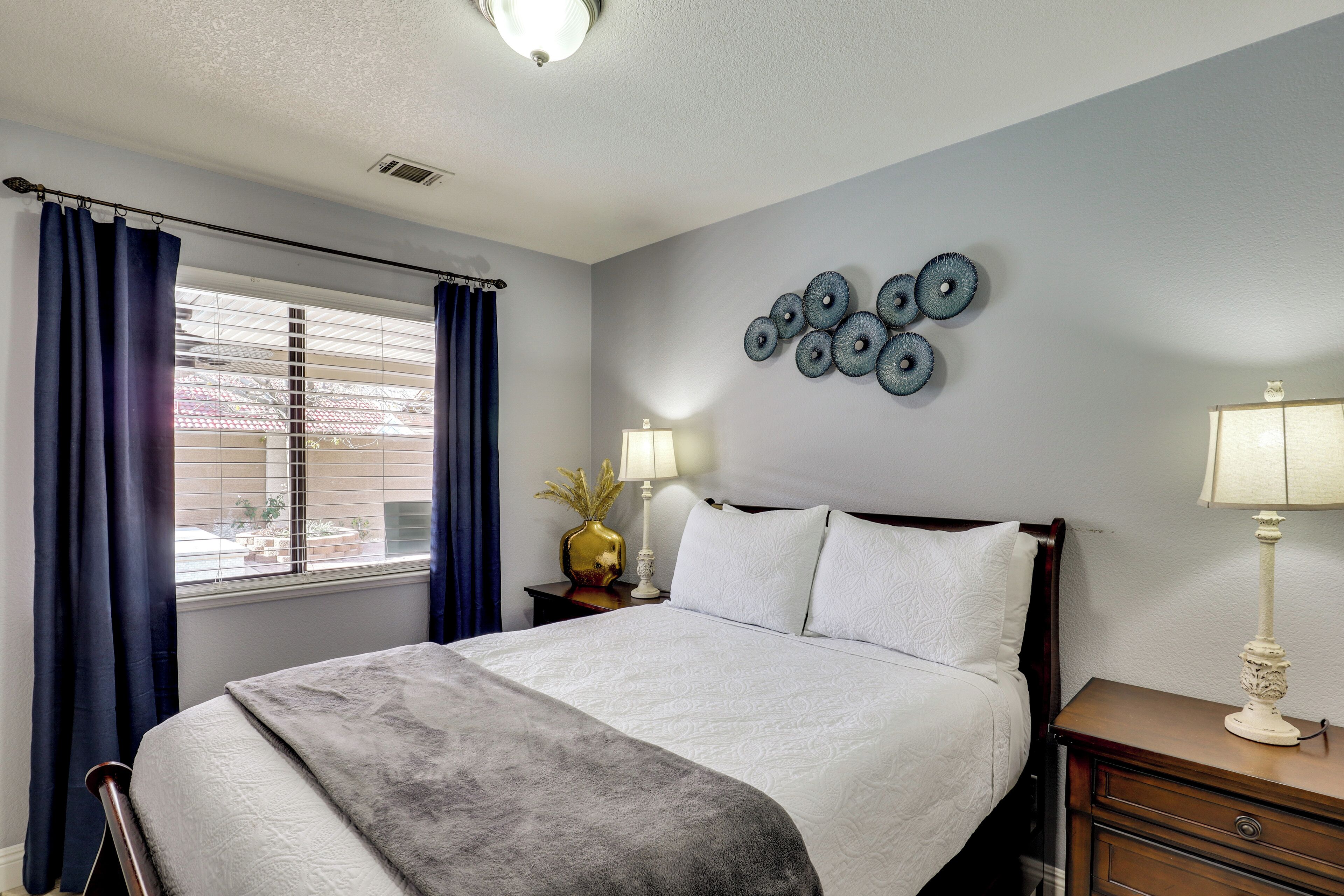 A neatly arranged hotel room with a large bed, a bedside table with a lamp, a wall clock, and a window with curtains.