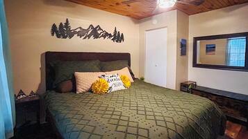 A bedroom with a bed, a headboard, a wall-mounted mirror, a door, and a small bedside table.