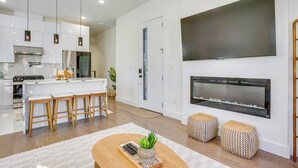 A modern kitchen with white cabinets, a central island with bar stools, a large flat-screen TV mounted above a fireplace, and a wooden coffee table with a plant. - Spacious & Upscale Family Retreat: 5 Mi to NYC (Jersey City)