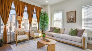 A living room with a sofa, armchair, and coffee table, a potted plant, and large windows with curtains. - Spacious & Upscale Family Retreat: 5 Mi to NYC (Jersey City)
