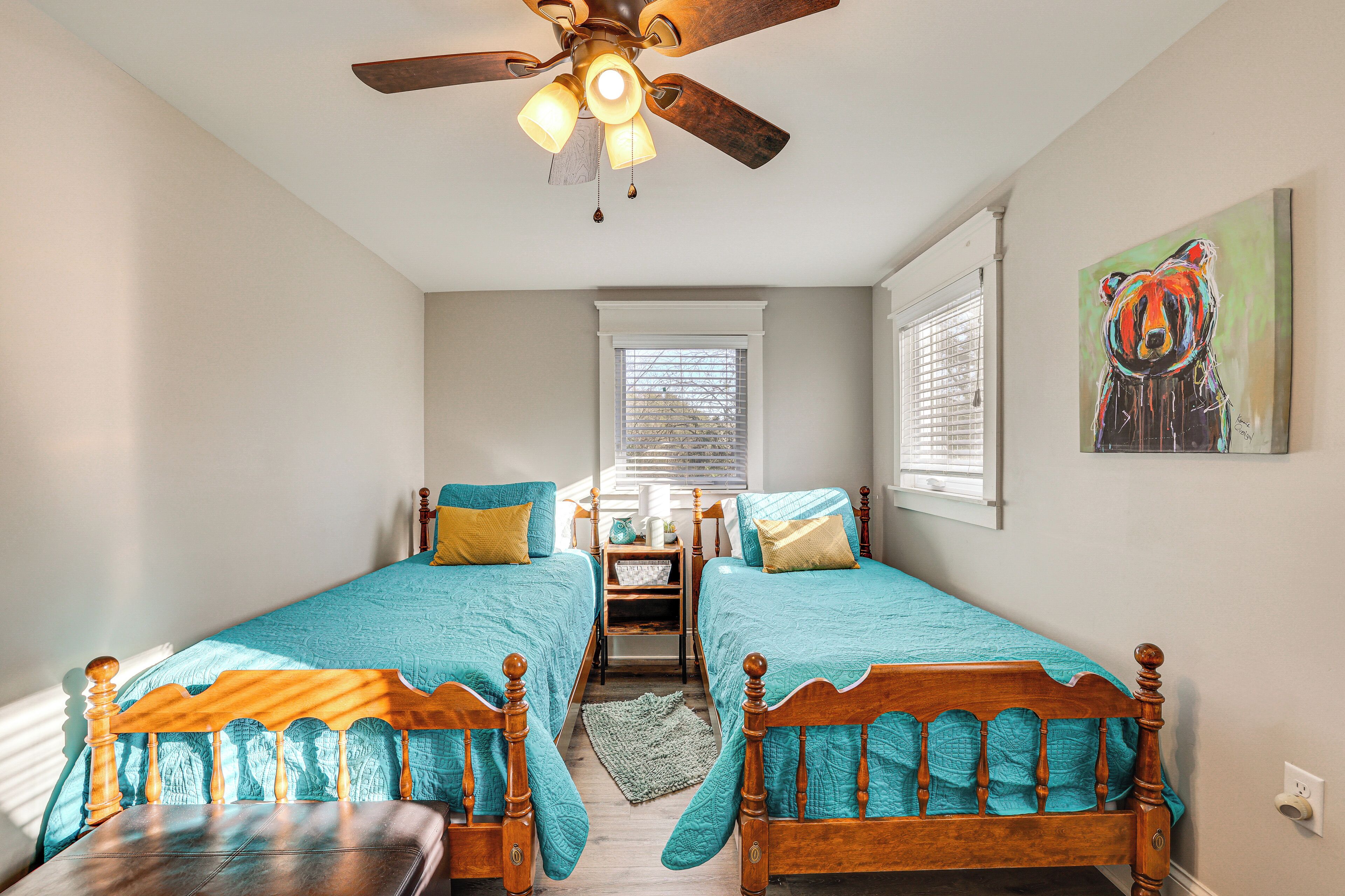 A bedroom with two wooden beds, a ceiling fan, a window with blinds, and a painting on the wall.