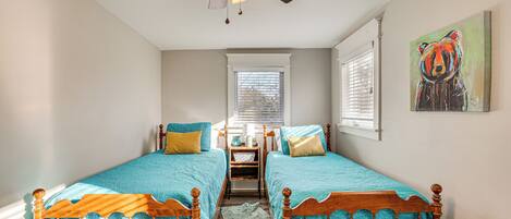 A bedroom with two wooden beds, a ceiling fan, a window with blinds, and a painting on the wall.