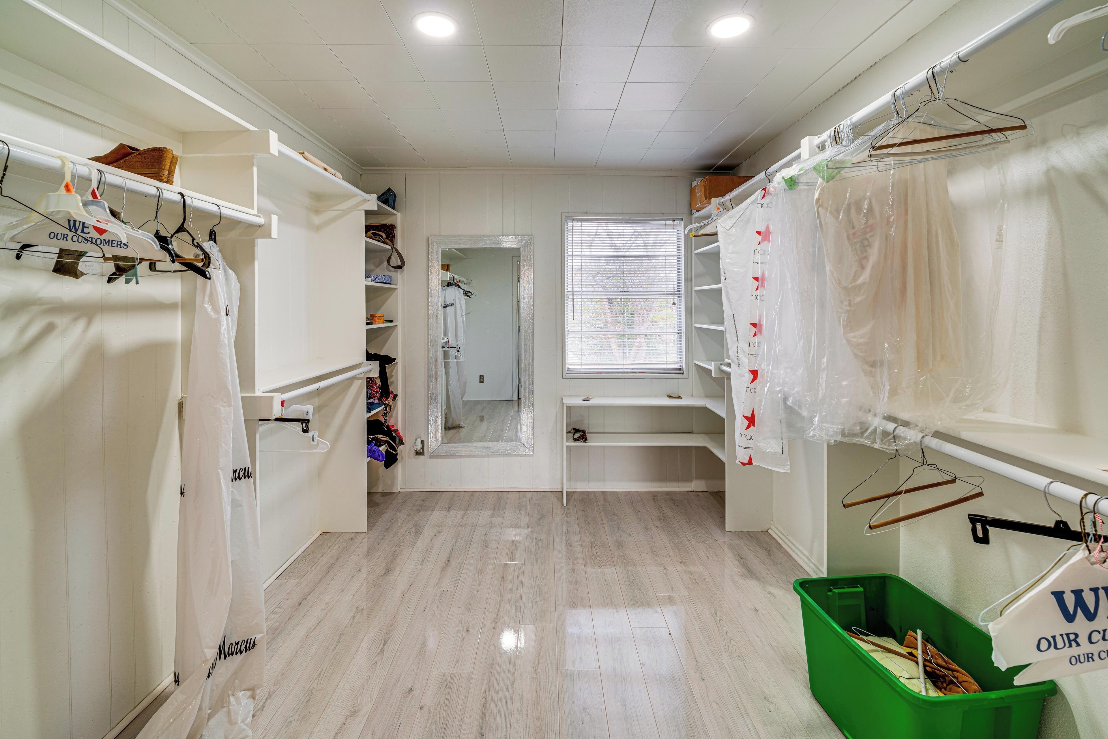 A hotel room with a wooden floor, a closet with hanging clothes, a window with blinds, and a green trash bin.