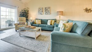 Un salon avec un canapé vert, un fauteuil à motifs floraux et une table basse. Deux œuvres d’art sont accrochées au mur, et une lampe sur pied est posée au sol.