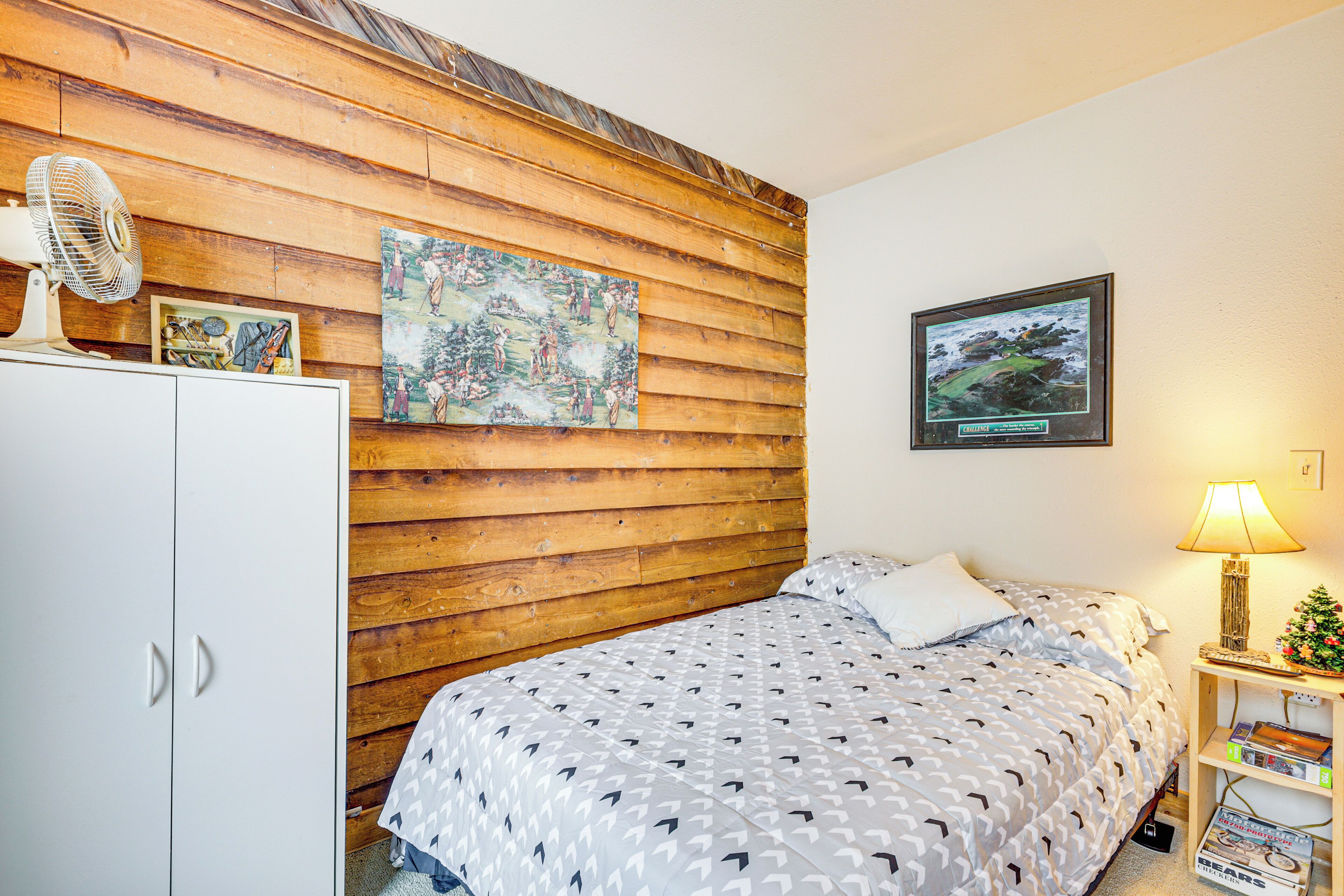 Une chambre avec une tête de lit en bois, un lit avec une literie à motifs, une armoire blanche, un ventilateur, un tableau encadré, une petite table avec une lampe et un sapin de Noël.