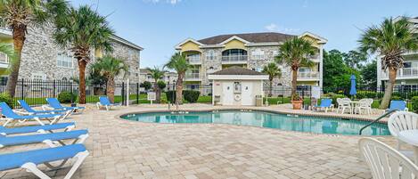 A pool area with lounge chairs, a pool, and a building with balconies and palm trees.
