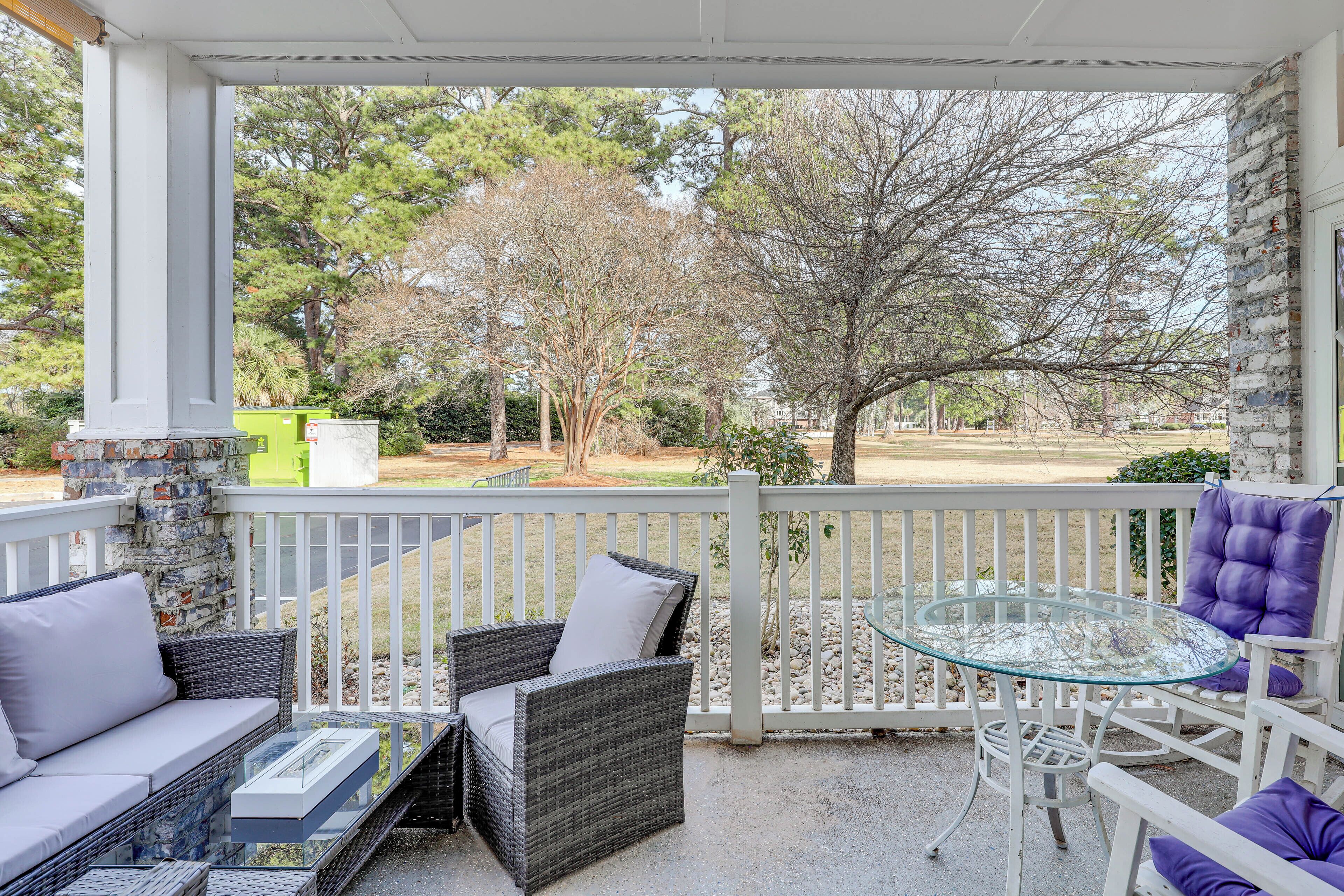 A covered outdoor seating area with wicker furniture and a glass table, overlooking a landscaped yard with trees and a building in the background.
