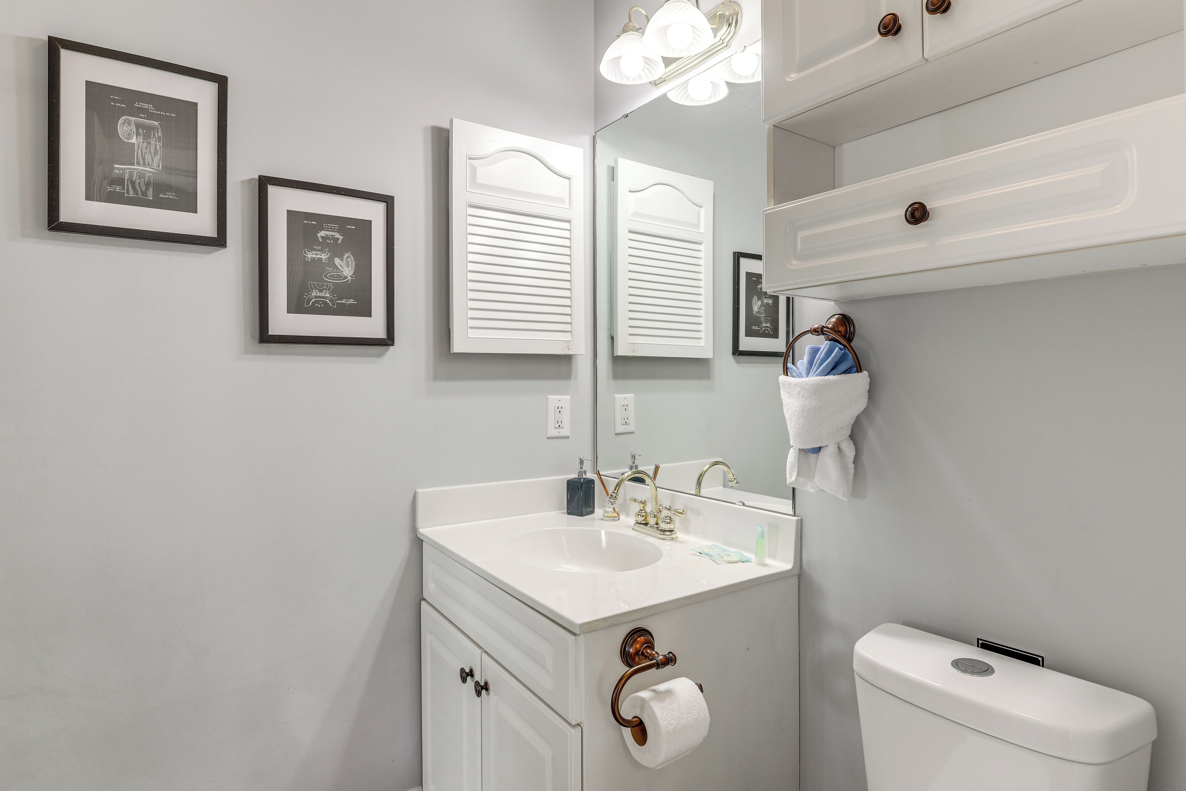 Een badkamer met een wit wastafelmeubel, toilet en een spiegel met verlichting erboven. Er hangen twee afbeeldingen in lijsten aan de muur.