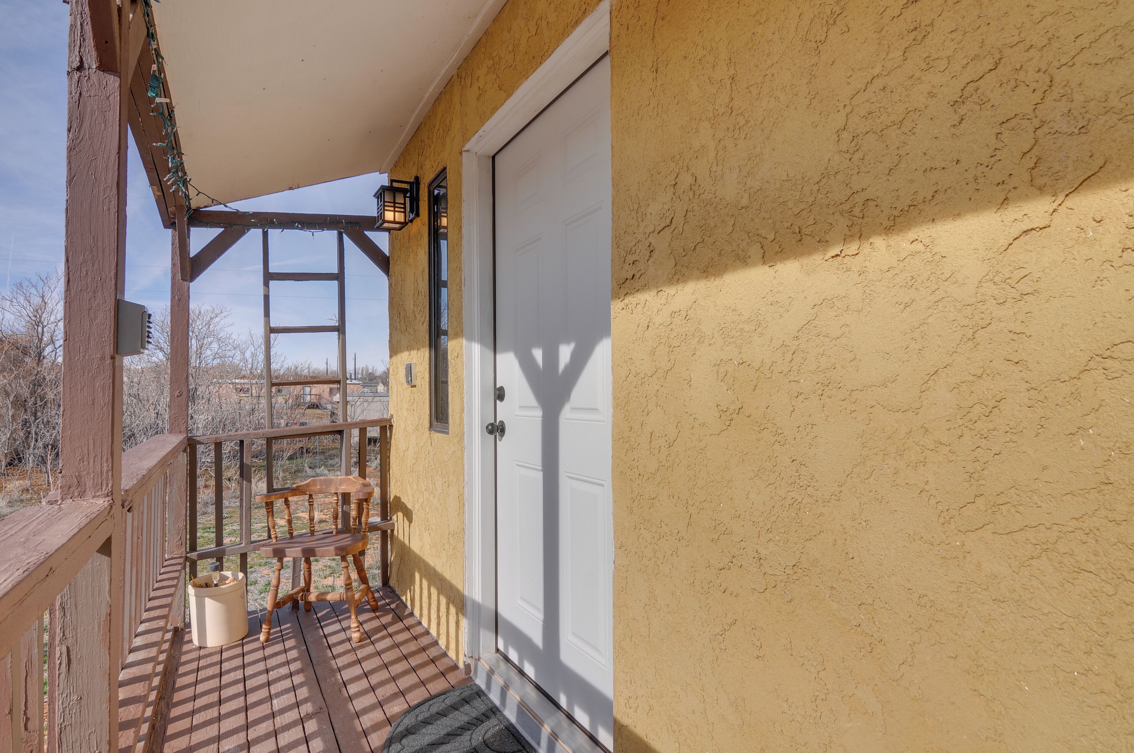 A wooden deck with a chair and a small table, a white door, and a ladder against a yellow wall.