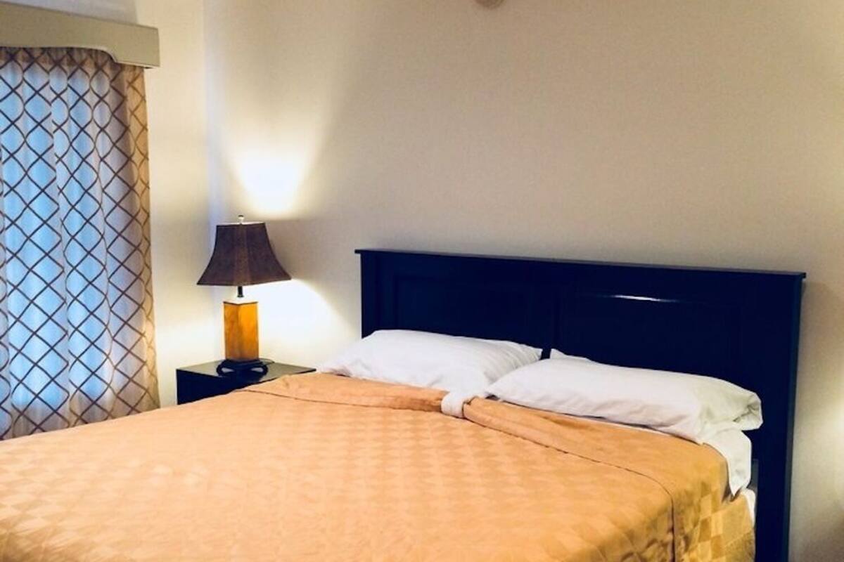 A neatly made bed with a brown quilt, a bedside lamp, and a window with patterned curtains.