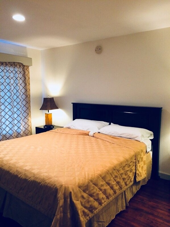 Una cama bien hecha con un edredón marrón, una lámpara de noche y una ventana con cortinas estampadas.
