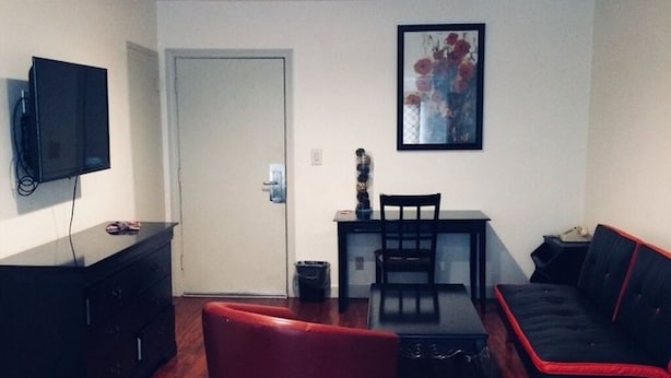 A living room with a wooden floor, a black sofa, a red chair, a black table, a TV on a stand, and a door.