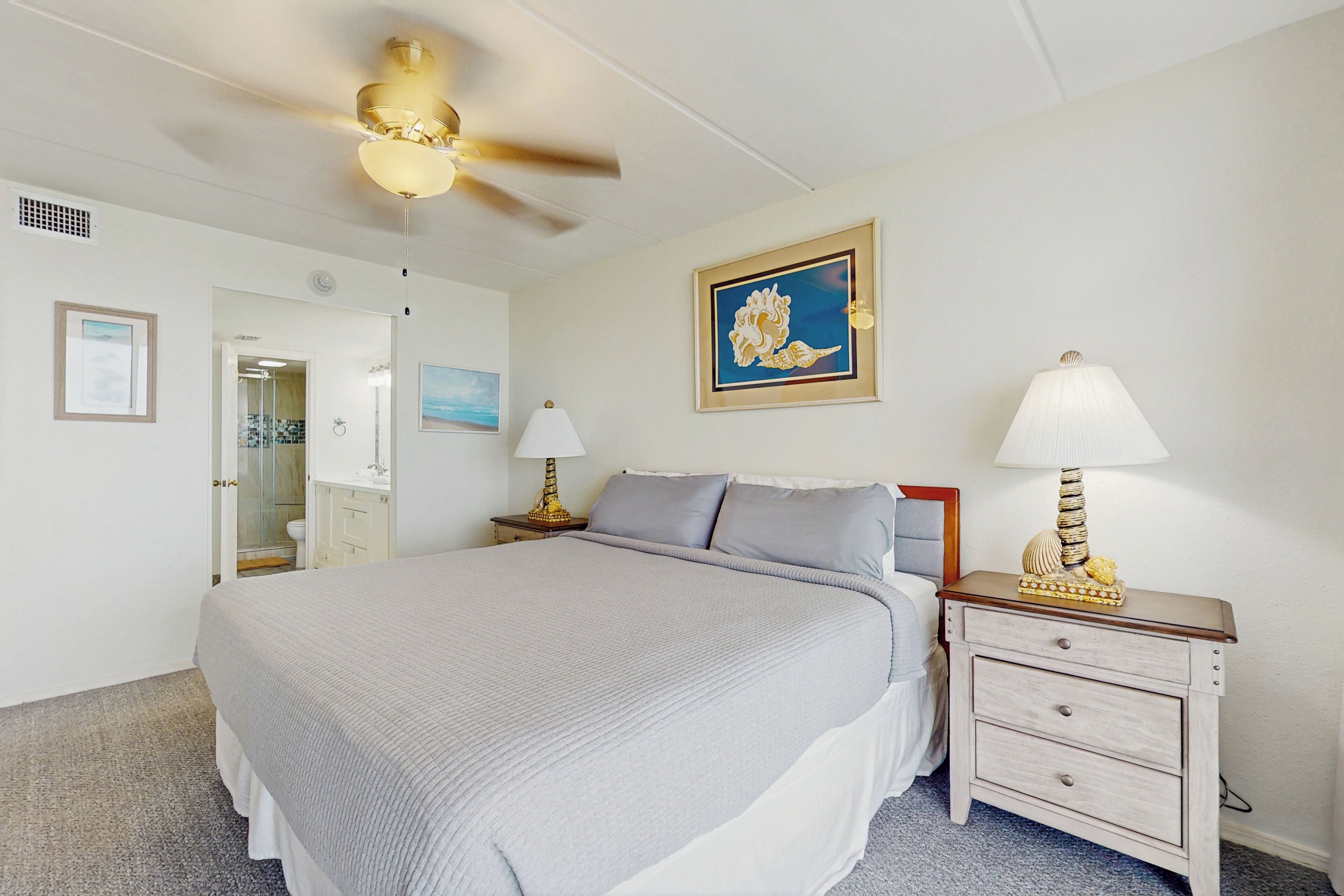 A bedroom with a bed, a nightstand with a lamp, a ceiling fan, and a framed picture on the wall.