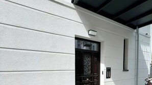 Ein weißes Gebäude mit einer dunkelholzigen Tür, einem kleinen Fenster und einer Überwachungskamera.