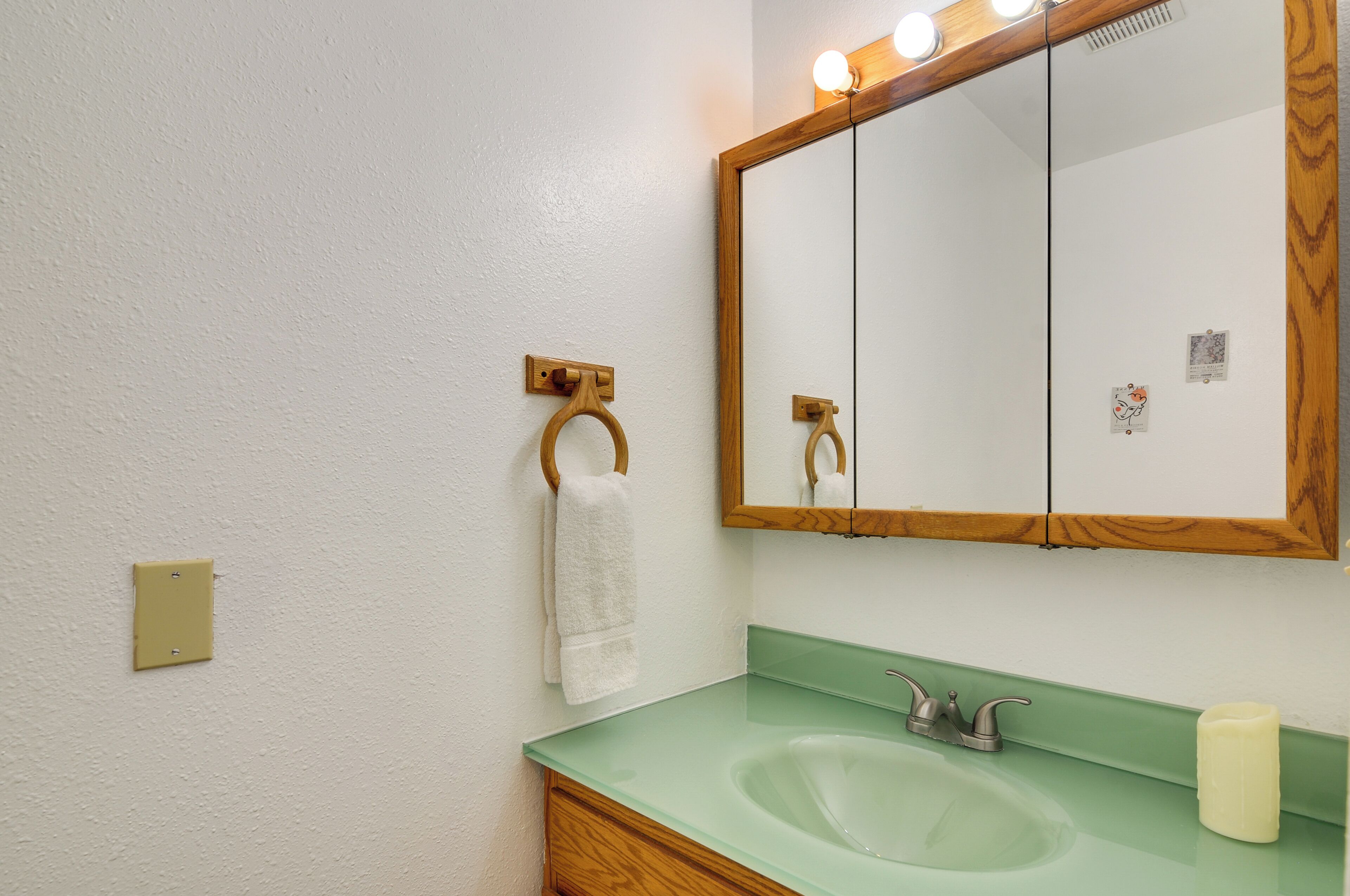 A bathroom with a green sink, a wooden towel rack, a mirror with lights, and a wall switch.