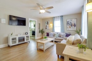A modern living room with a sofa, coffee table, and TV. There is a ceiling fan and a dining table with a plant. - Walk to Beach! Norfolk Family Home w/ Yard (Norfolk)