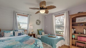 Une chambre avec deux lits, un ventilateur de plafond, une fenêtre avec des rideaux, une bibliothèque et une vue sur l’extérieur.