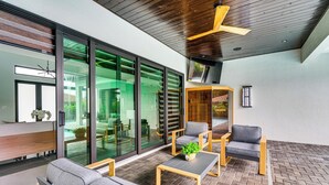 A modern outdoor seating area with a wooden ceiling, a TV mounted on the wall, and a glass door leading to an interior space. - Luxe Outdoor Space! Pet-friendly Cape Coral Home (Cape Coral)