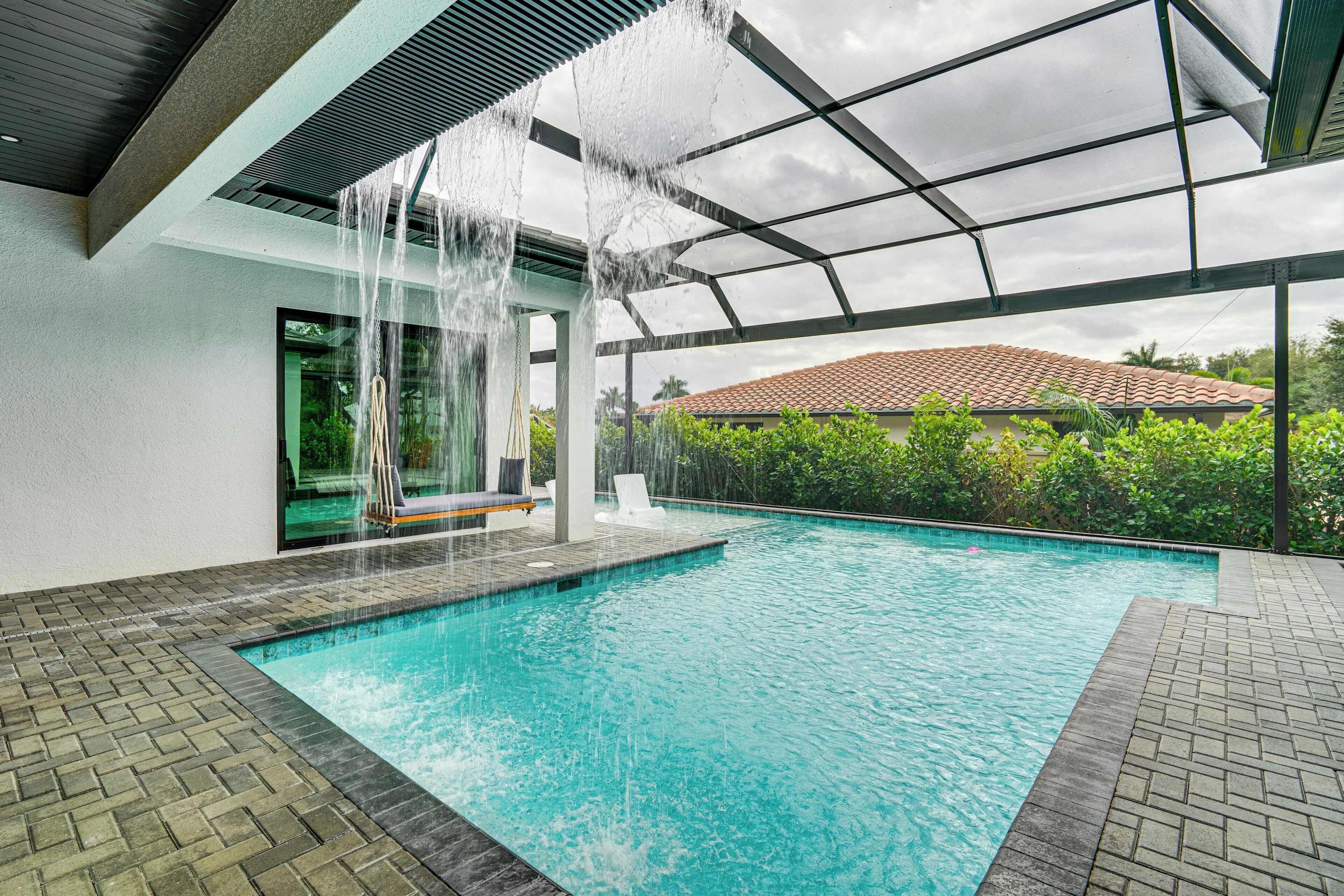 A swimming pool with a waterfall feature, surrounded by a covered area with a view of a red-tiled roof and greenery.