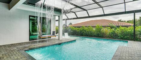 A swimming pool with a waterfall feature, surrounded by a covered area with a view of a red-tiled roof and greenery.