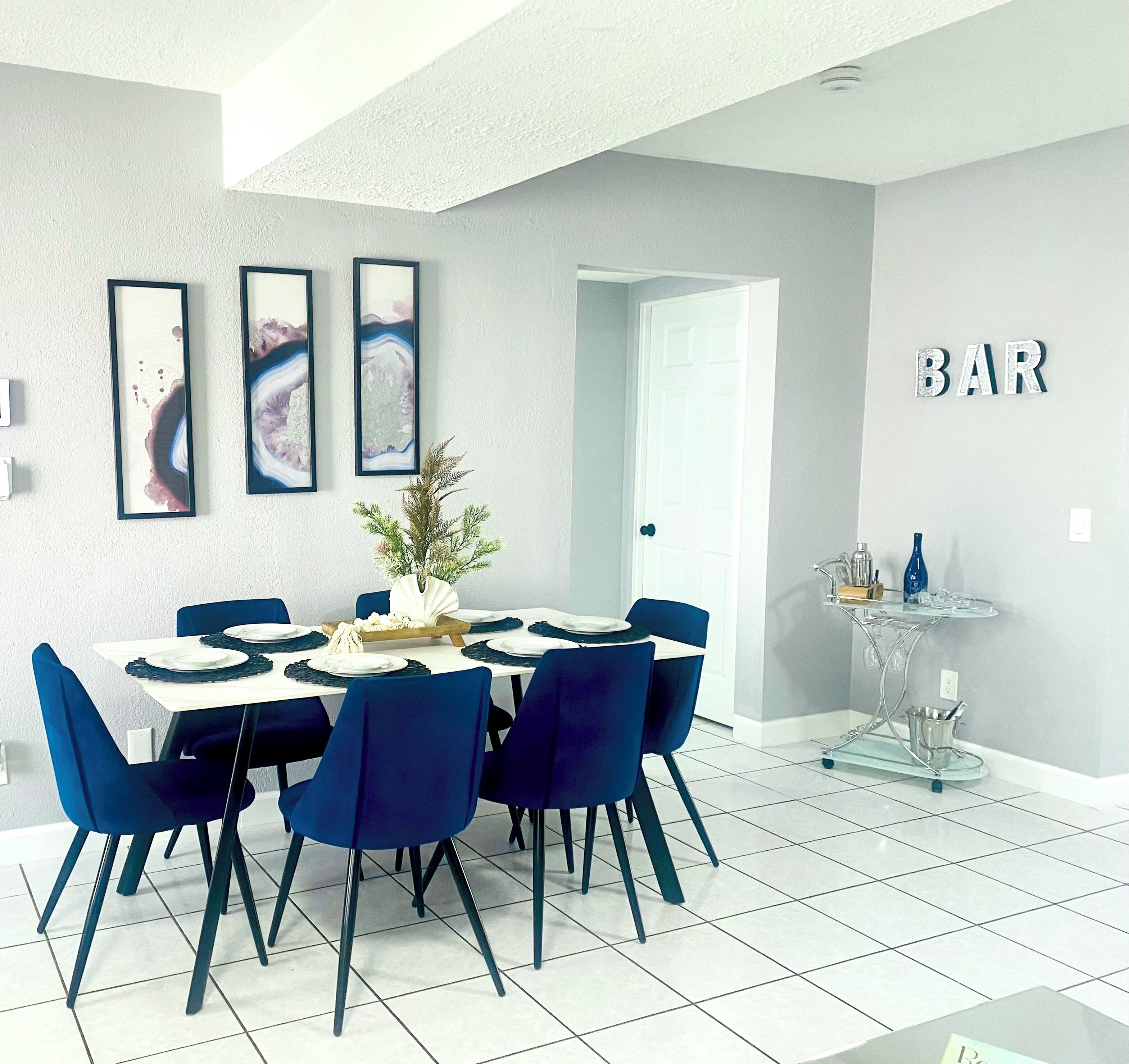 Un espace repas moderne avec une table blanche dressée pour six personnes, des chaises bleues et trois œuvres d’art encadrées accrochées au mur.