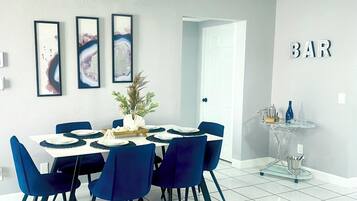 Un espace repas moderne avec une table blanche dressée pour six personnes, des chaises bleues et trois œuvres d’art encadrées accrochées au mur.
