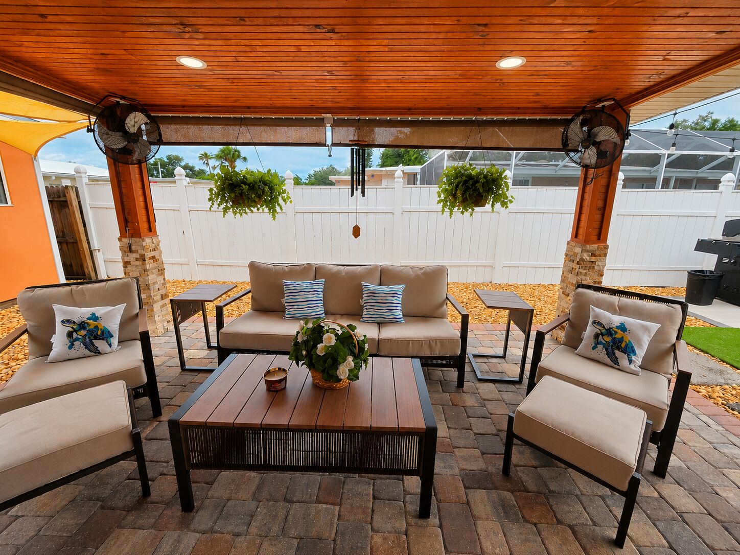 An outdoor patio with a wooden ceiling, beige sofa, two armchairs, a coffee table, and hanging plants.