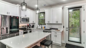 A modern kitchen with white cabinets, stainless steel appliances, a large island with bar stools, and a door with a window with blinds. - Treehouse Escape in Tahitian Village (Bastrop)