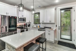 A modern kitchen with white cabinets, stainless steel appliances, a large island with bar stools, and a door with a window with blinds. - Treehouse Escape in Tahitian Village (Bastrop)
